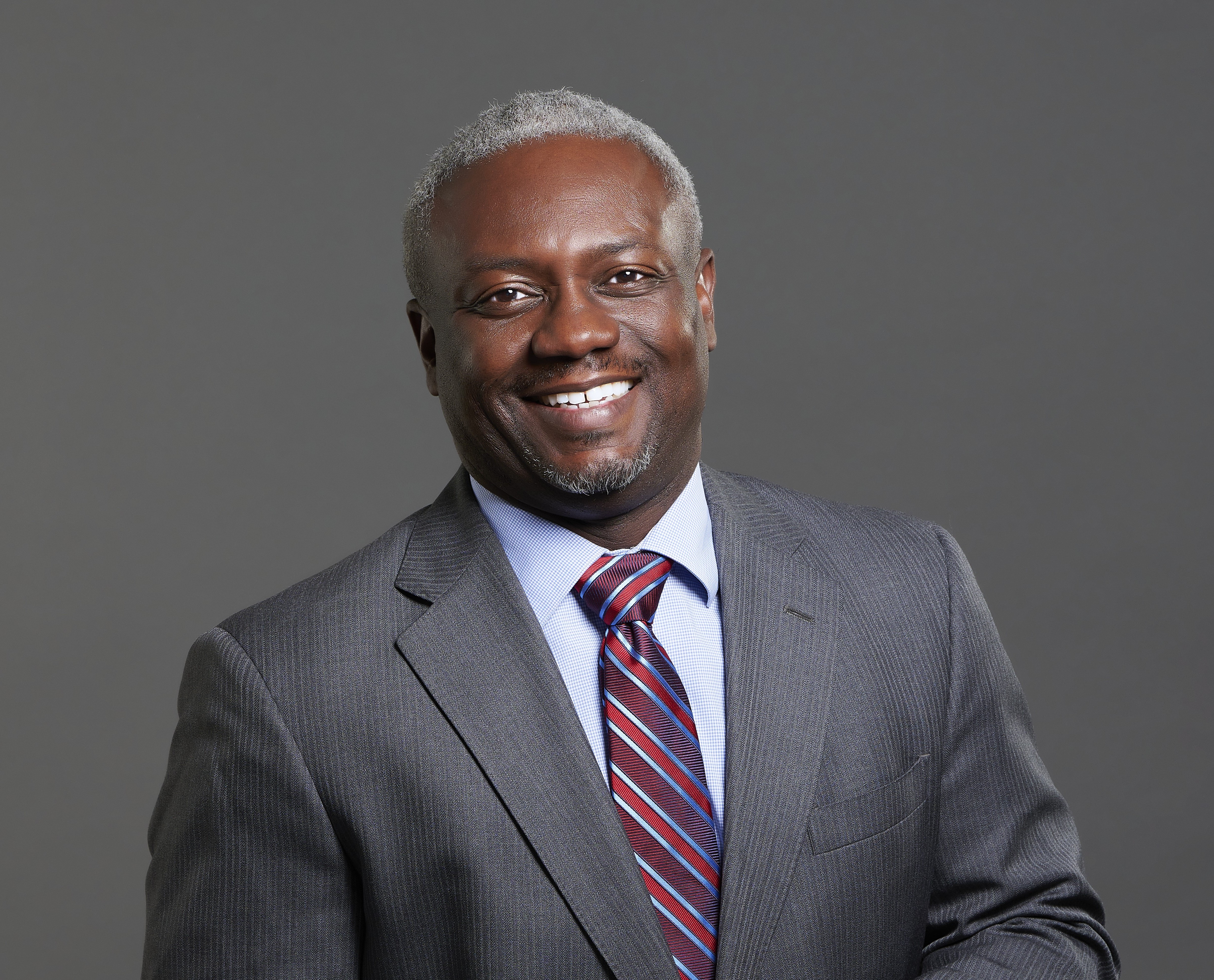 Black man with grey hear smiling in a grey suit and stripped tie